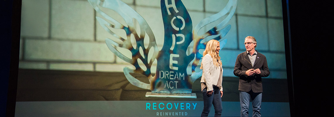 Governor Doug Burgum, wearing a black suit coat and jeans, stands to the right of First Lady Kathryn Burgum, wearing a white jacket and jeans, in front of a screen with a picture of an award that says Hope, Dream, Act.