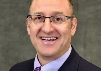 Man wearing glasses and suit jacket with purple shirt and tie smiles